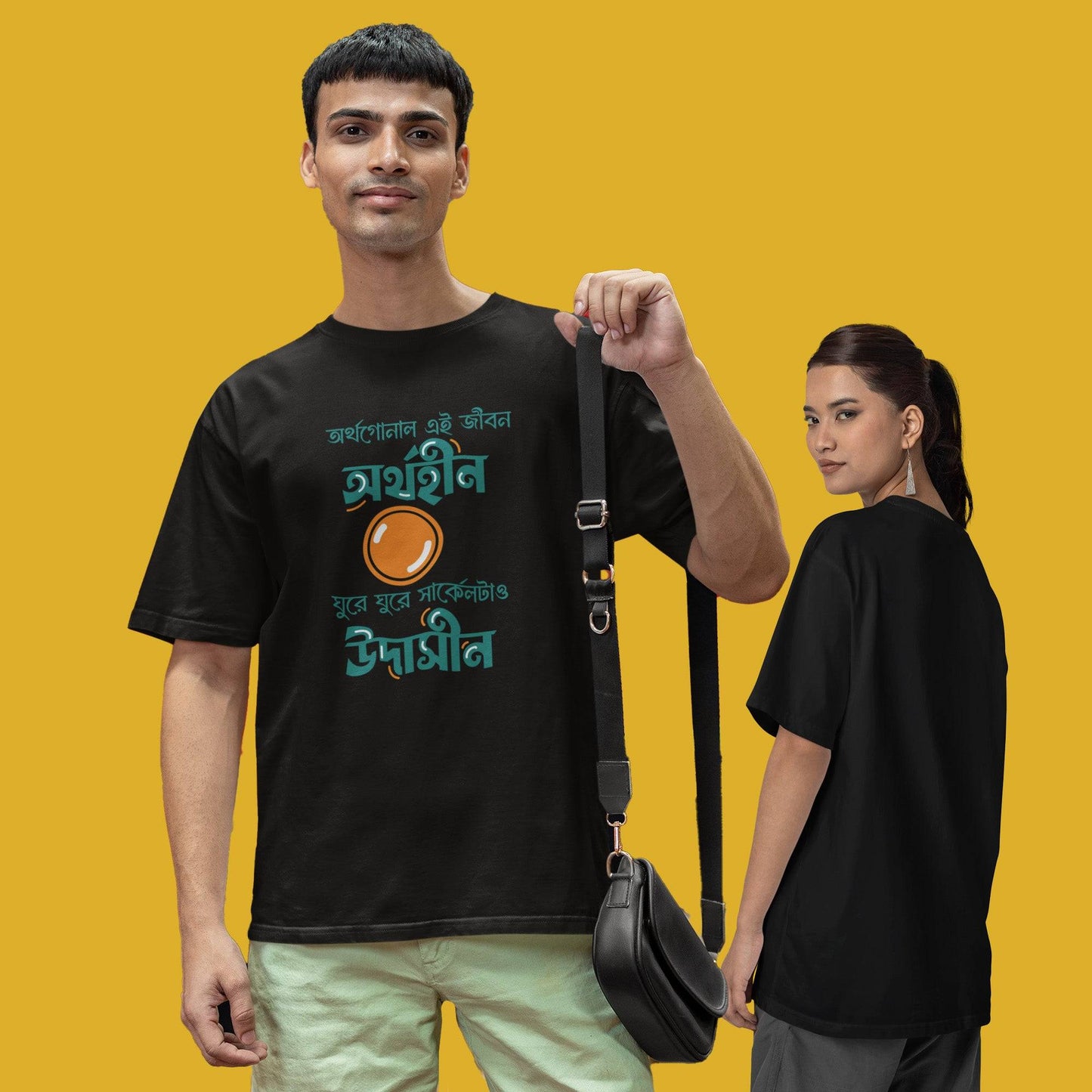 Black oversized unisex T-shirt with funny Bengali typography caption, modeled by a man and woman against a yellow background.