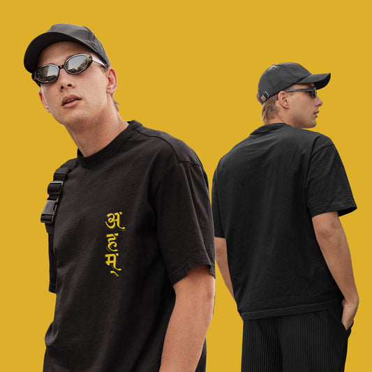 Model wearing Smarteez's black oversized unisex T-shirt 'Ahm,' featuring yellow Sanskrit typography, styled with a cap and casual accessories.