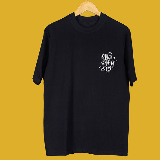 Black oversized unisex T-shirt with 'Ami Amar Moto' Bengali typography in white, hanging on a wooden hanger against a yellow background.