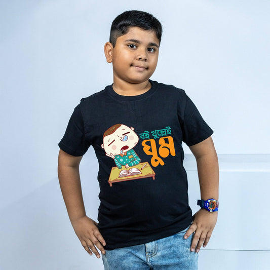 Bengali TShirt for kids featuring 'Boi Khulle i Ghum' text and a cartoon of a sleepy boy at a desk. Black cotton T-shirt, soft and breathable.