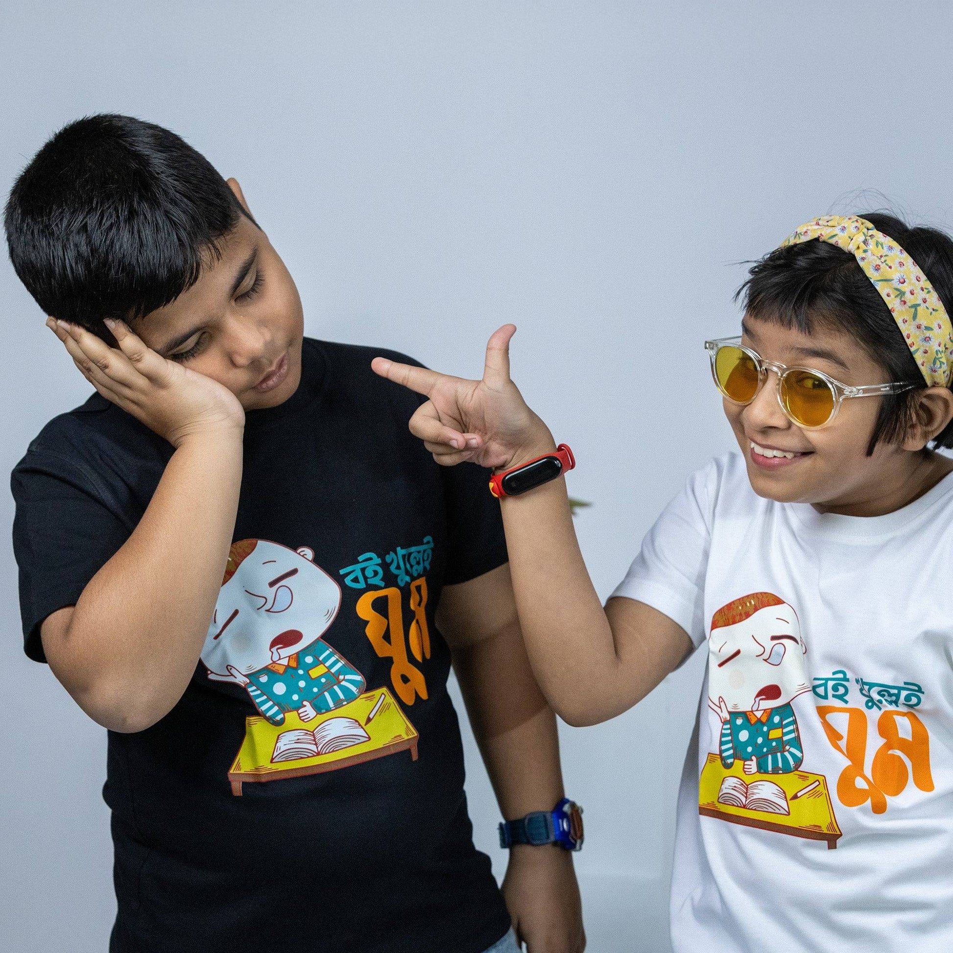 Two kids wearing 'Boi Khulle i Ghum' Bengali TShirts, one in black and one in white, featuring a funny cartoon design. Perfect for kids' casual wear.